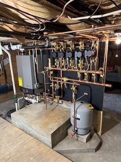 Utility room with boilers, copper pipes, and a water heater under exposed basement joists