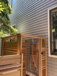 Wooden outdoor shower stall beside a house, with a hose and hanging showerhead.