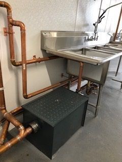 Industrial utility sink with exposed copper pipes and a gray metal box on the floor in a basement.