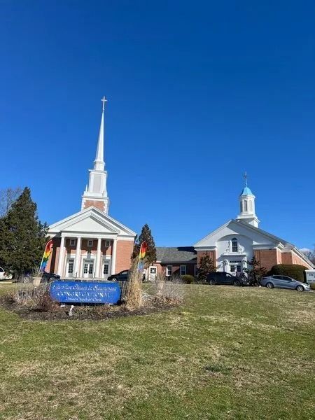 Two small white church buildings with steeples and rainbow flags on a grassy lawn under a blue sky