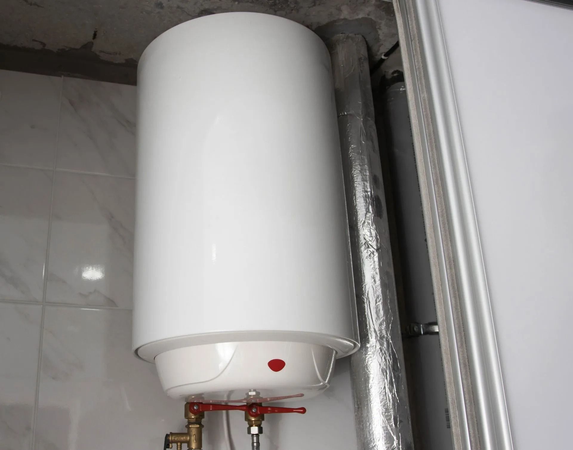 White cylindrical water heater mounted on a tiled wall with exposed pipes underneath.