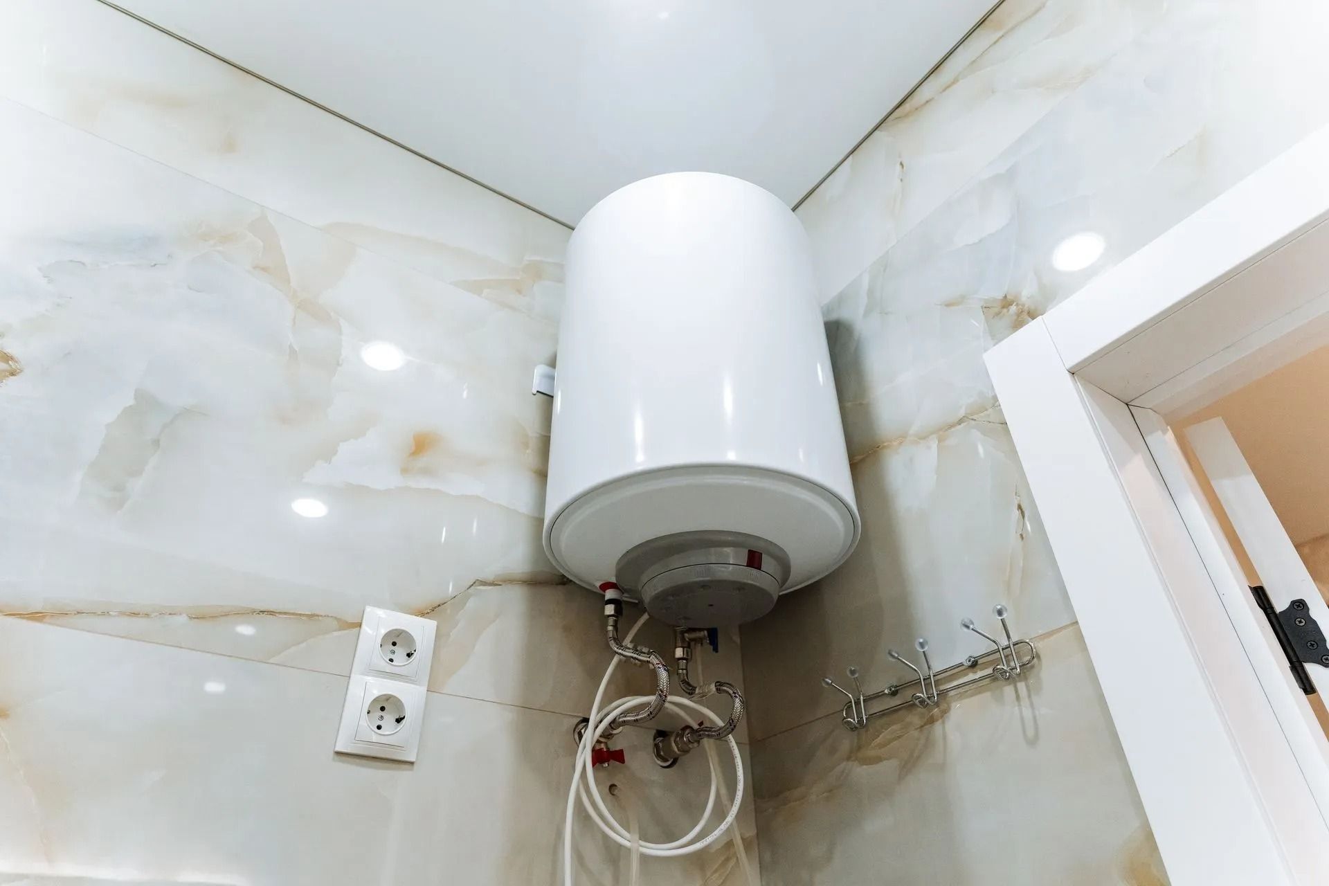 White wall-mounted water heater in a tiled bathroom corner, with exposed pipes and electrical outlet nearby