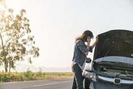 Picture of a lady on the phone leaning on her car with the hood open and waiting for a tow truck.