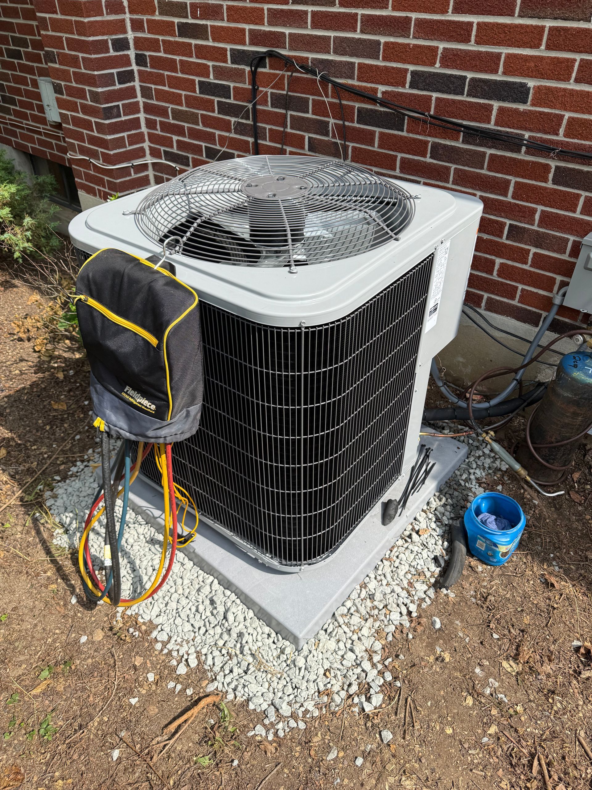 Outdoor air conditioner unit beside a brick house in a narrow grassy side yard.