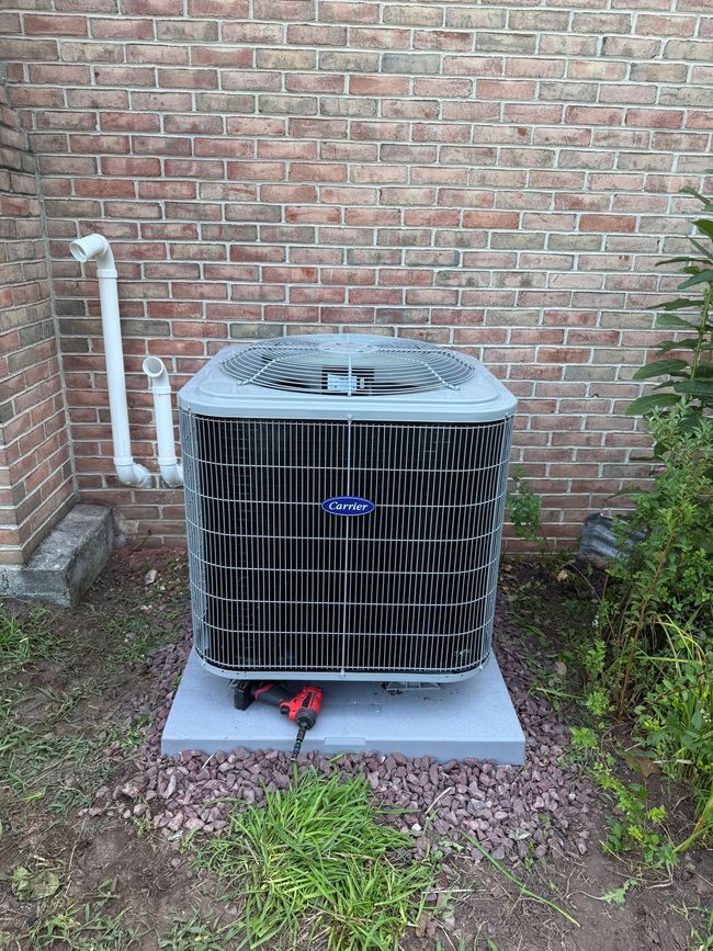 Outdoor air conditioner unit on a concrete pad beside a brick wall and shrubs