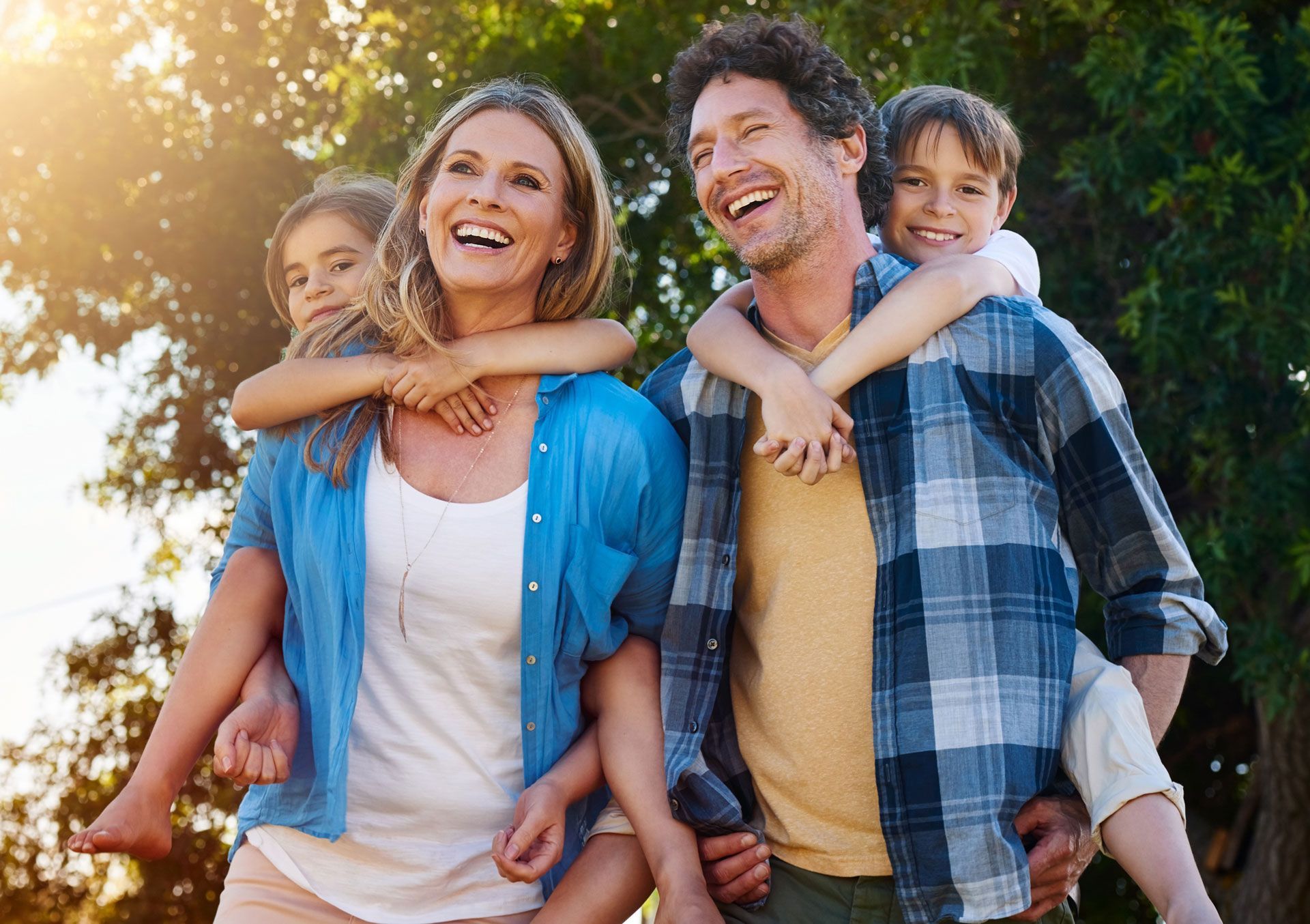 Smiling family outdoors in sunlight, with parents carrying two children on their backs.