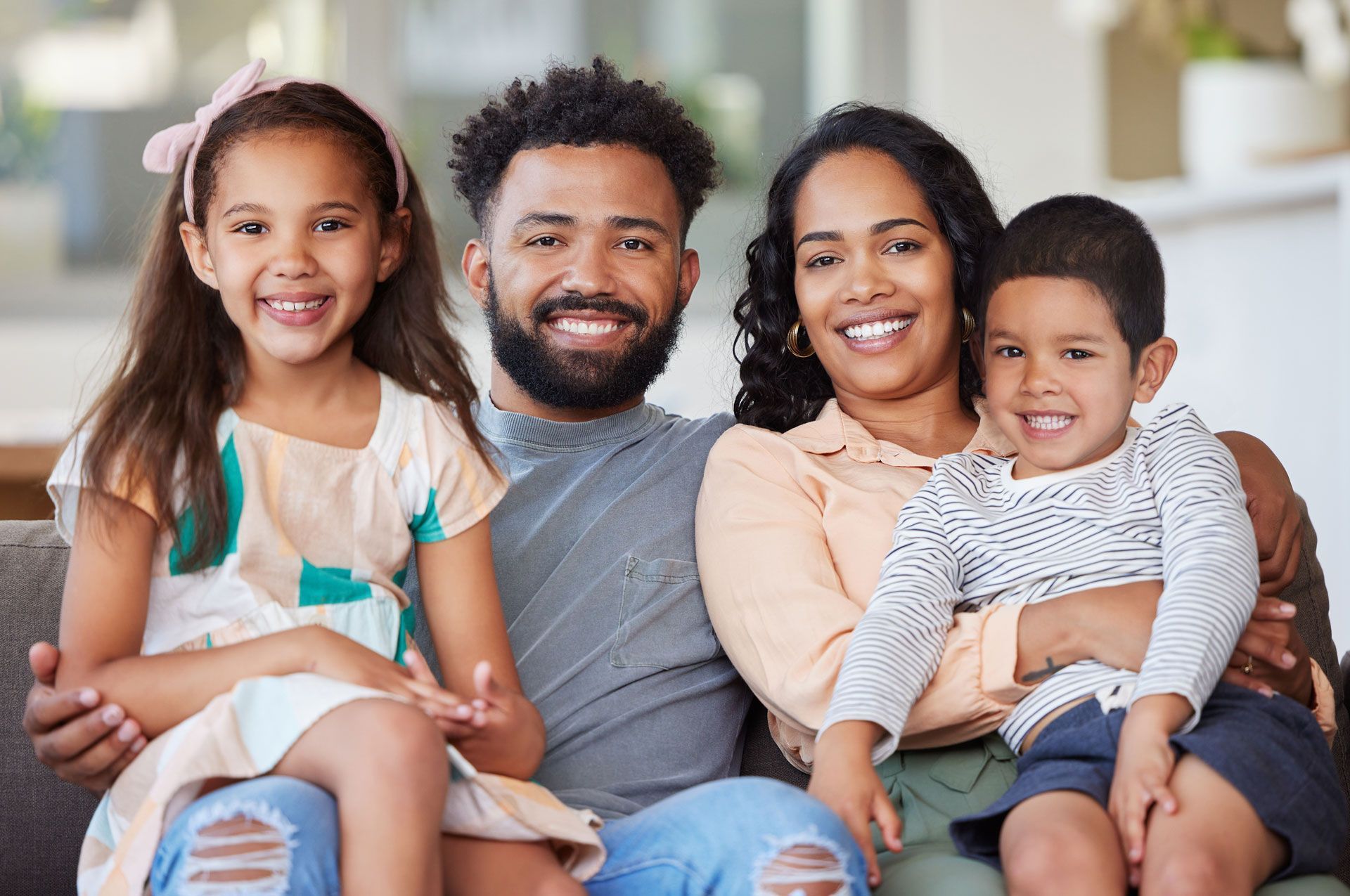 A smiling family of four sits together on a couch in a bright living room.