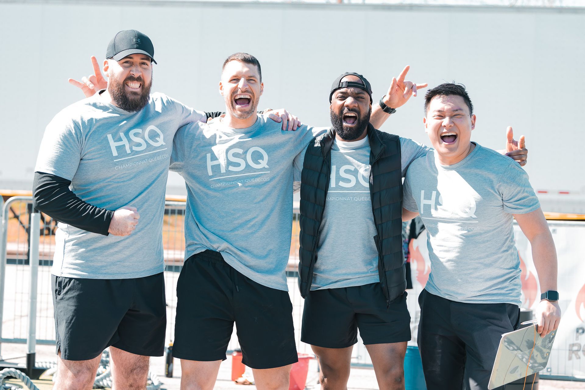 Quatre personnes vêtues de t-shirts gris HSO sourient et posent en extérieur, bras dessus bras dessous.