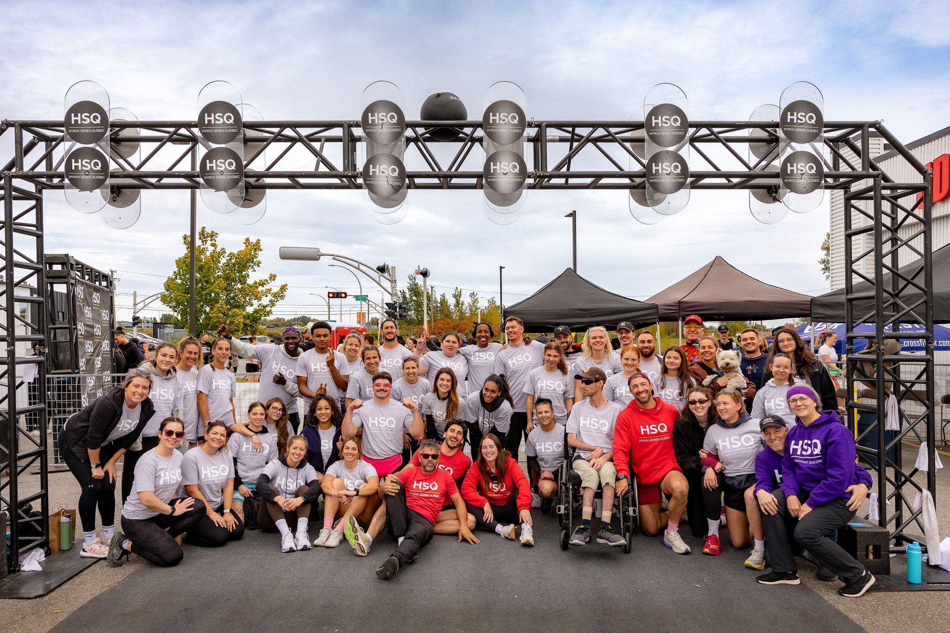 Group photo of runners and volunteers at a race finish line under a metal arch, with tents and lights behind them