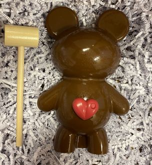 Brown gummy bear with a pink heart on its belly next to a small wooden mallet on a gray textured surface