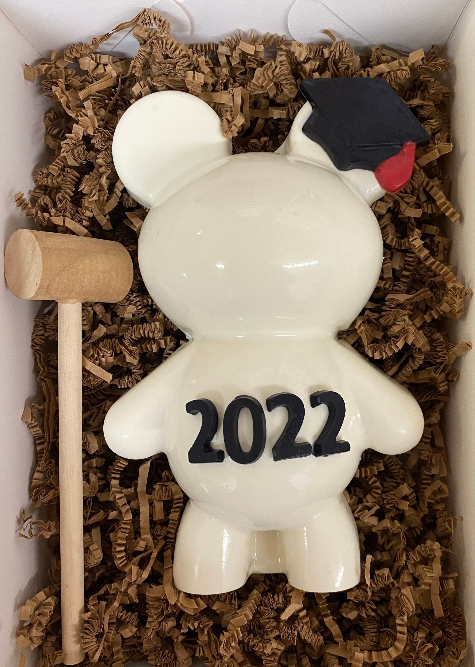 White ornament with “2022” on back, wearing black cap with red accent, in a box with shredded filler