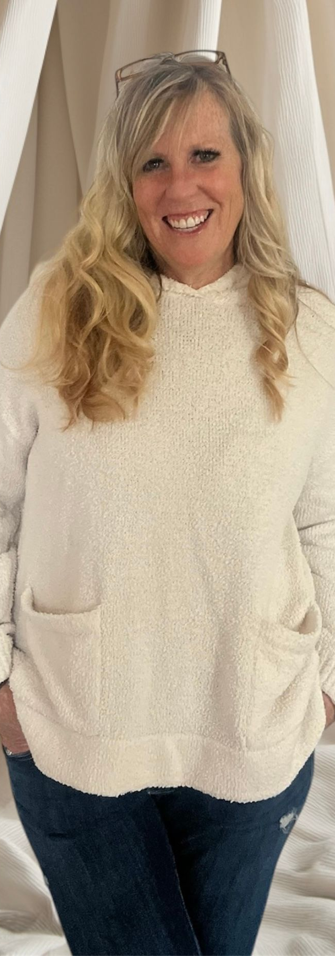 Woman standing indoors in a cream sweater and jeans, smiling at the camera.