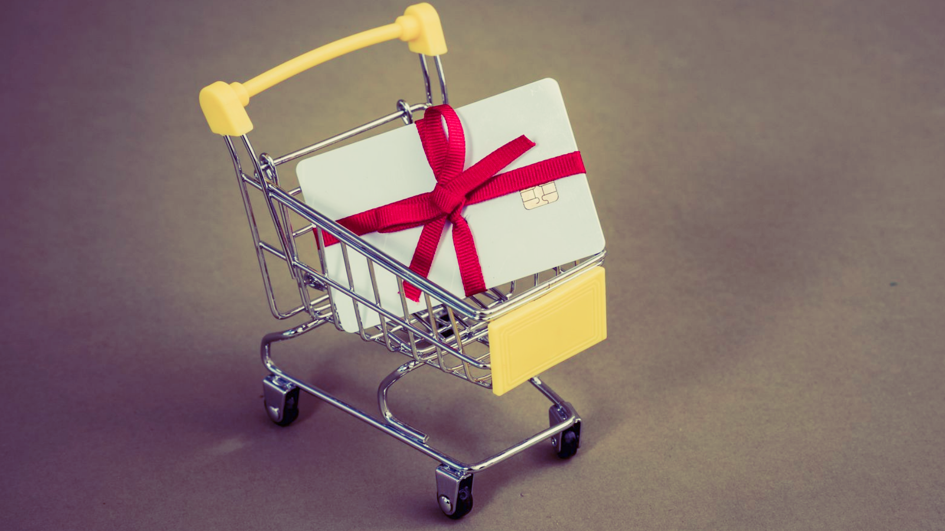 Mini shopping cart holding a white gift box wrapped with a red ribbon on a gray background
