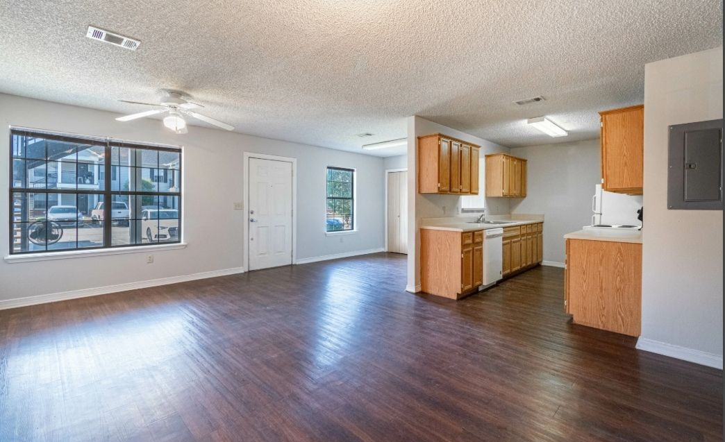 Empty open-plan apartment living area with dark wood floors, large windows, and a small kitchen.