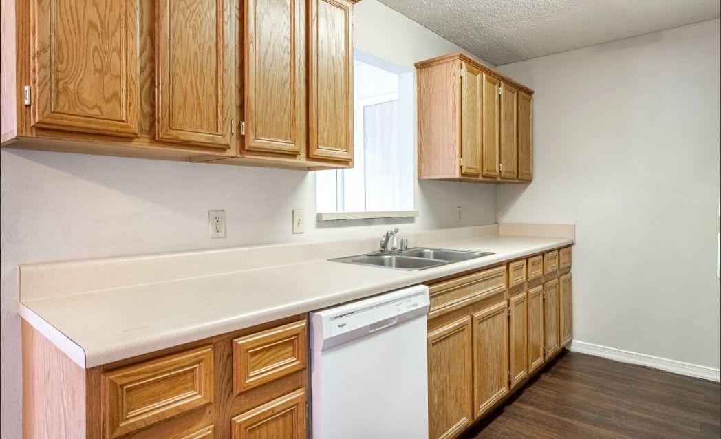 Bright kitchen with wooden cabinets, white countertops, sink, dishwasher, and window above the sink.