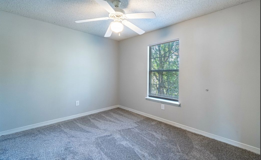 Empty light-gray bedroom with a ceiling fan and a window showing greenery