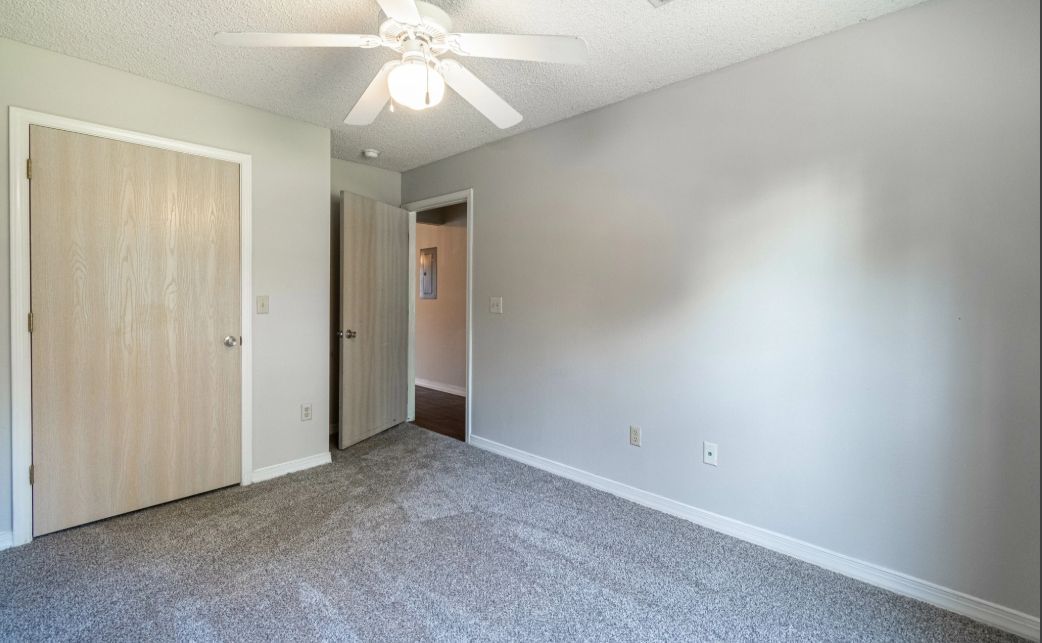 Empty bedroom with gray carpet, white walls, ceiling fan, and closet doors.