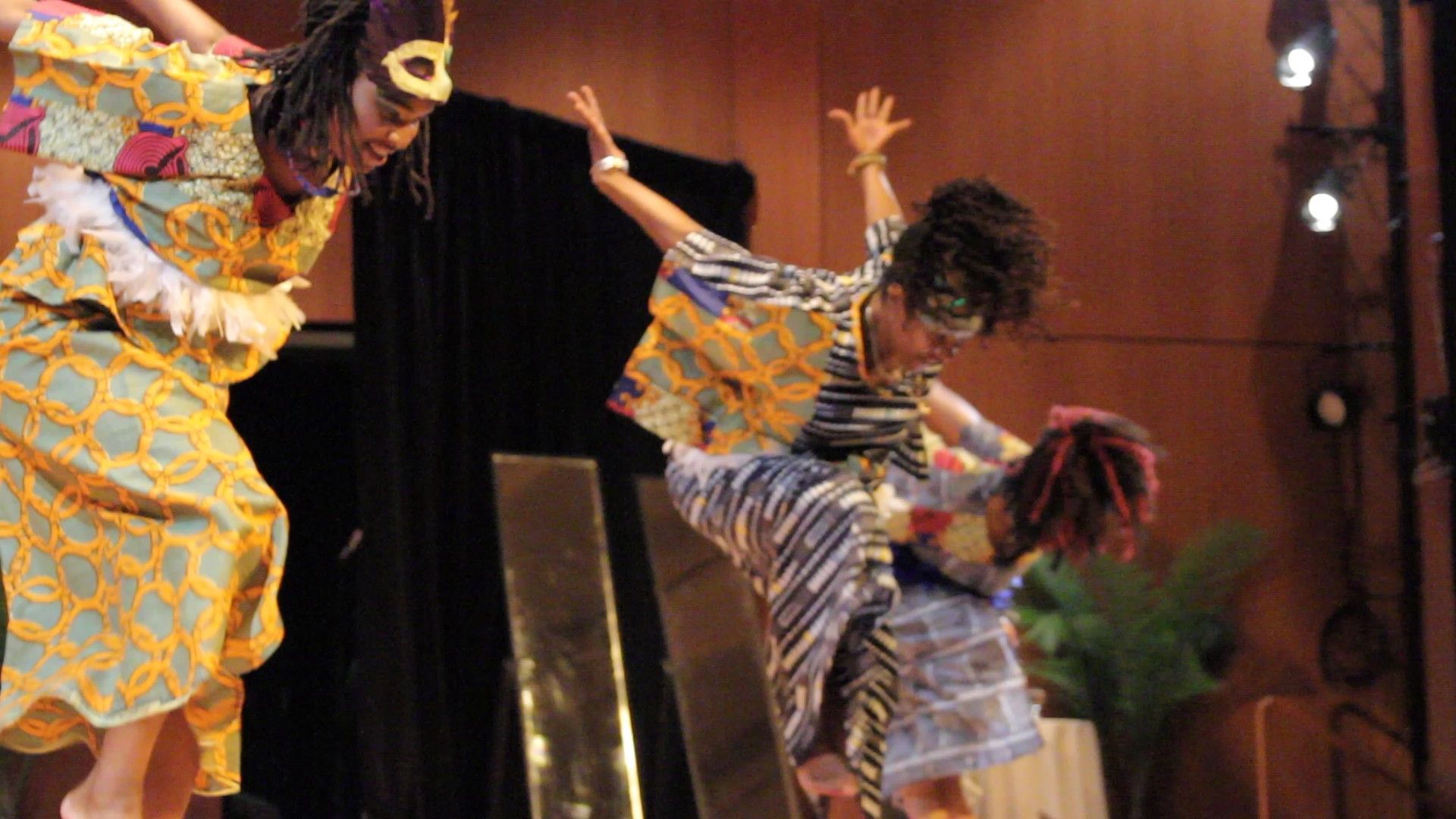Three dancers in vibrant costumes perform on stage, arms raised, bent at waist.