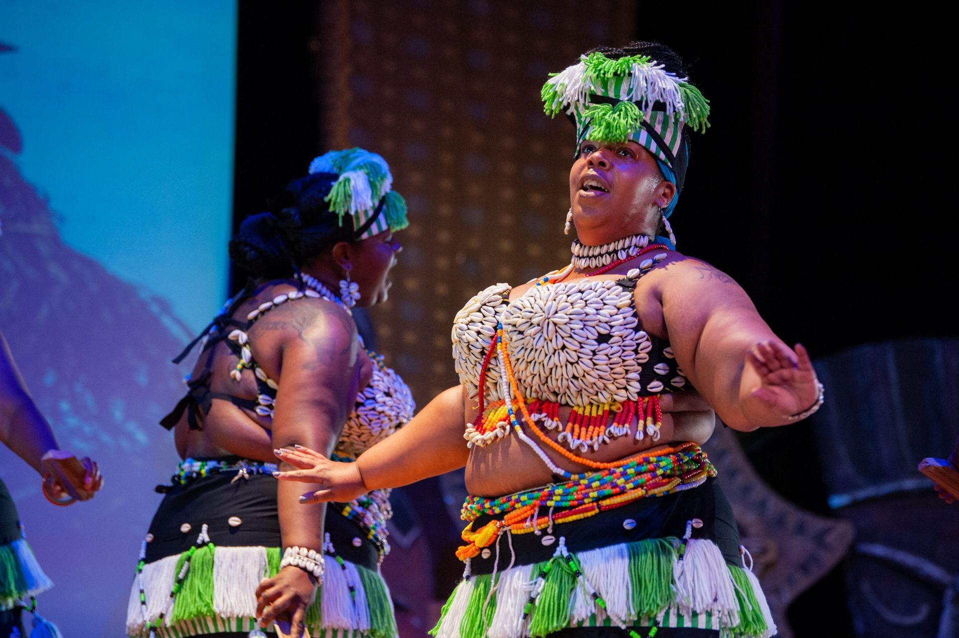 Women in traditional African clothing dance on stage, black and white skirts, beaded tops and headbands.
