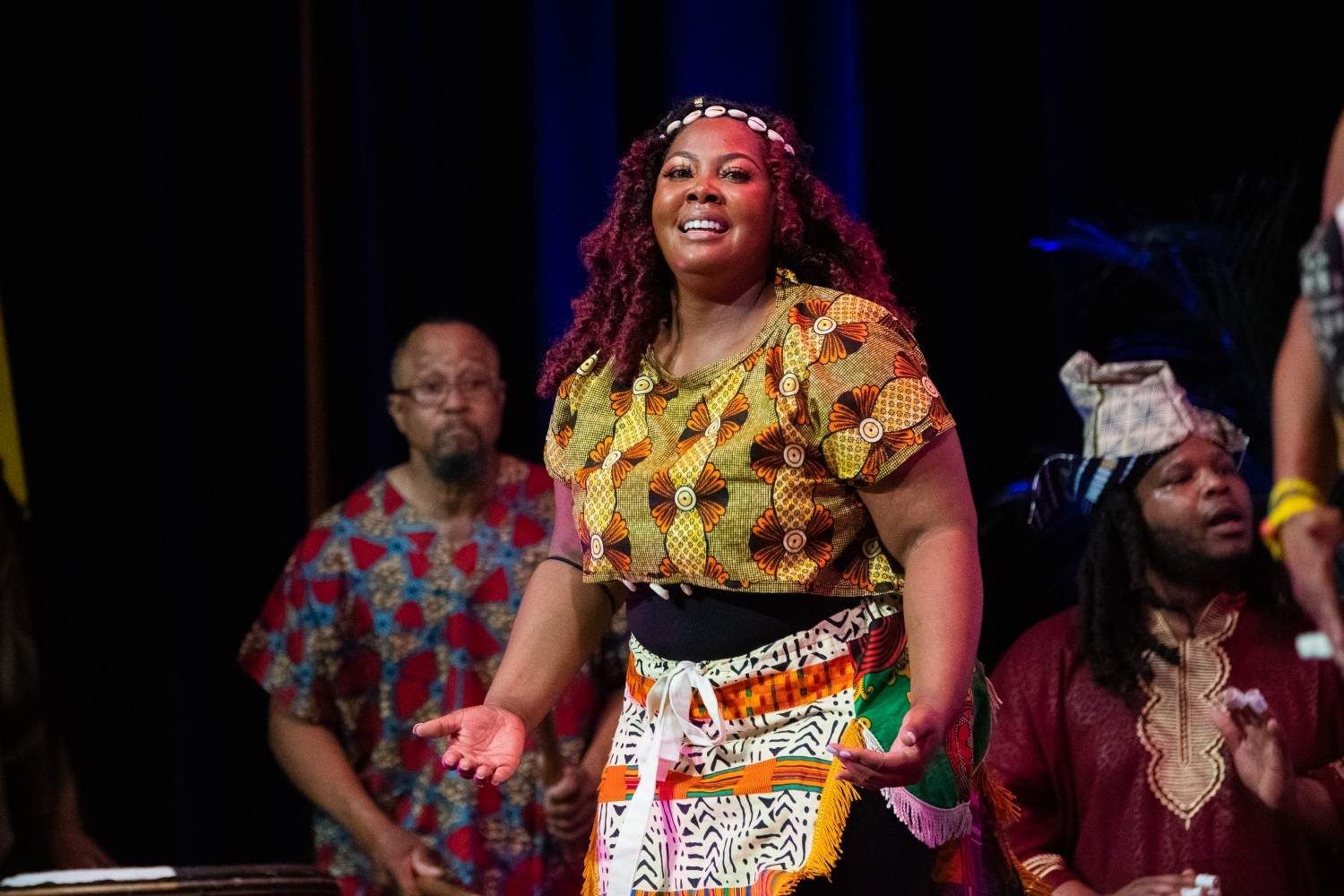 Woman in colorful African print outfit smiles, gesturing on stage. People with traditional attire in background.
