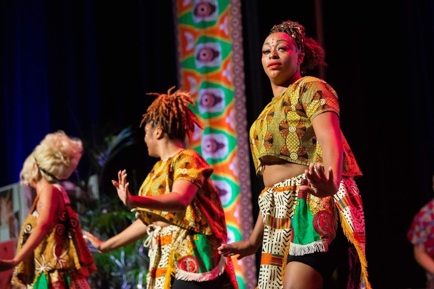 Three women in colorful African-print outfits dance on a stage, orange and green decor in the background.