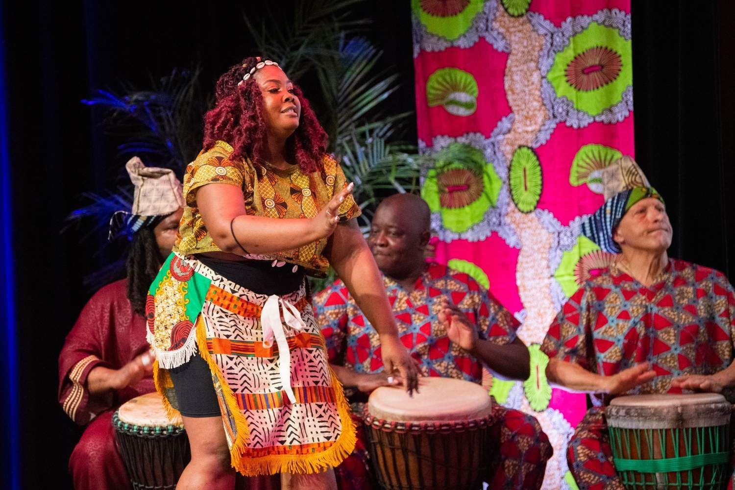 Woman dancing onstage, with drummers in traditional African attire, colorful backdrop.
