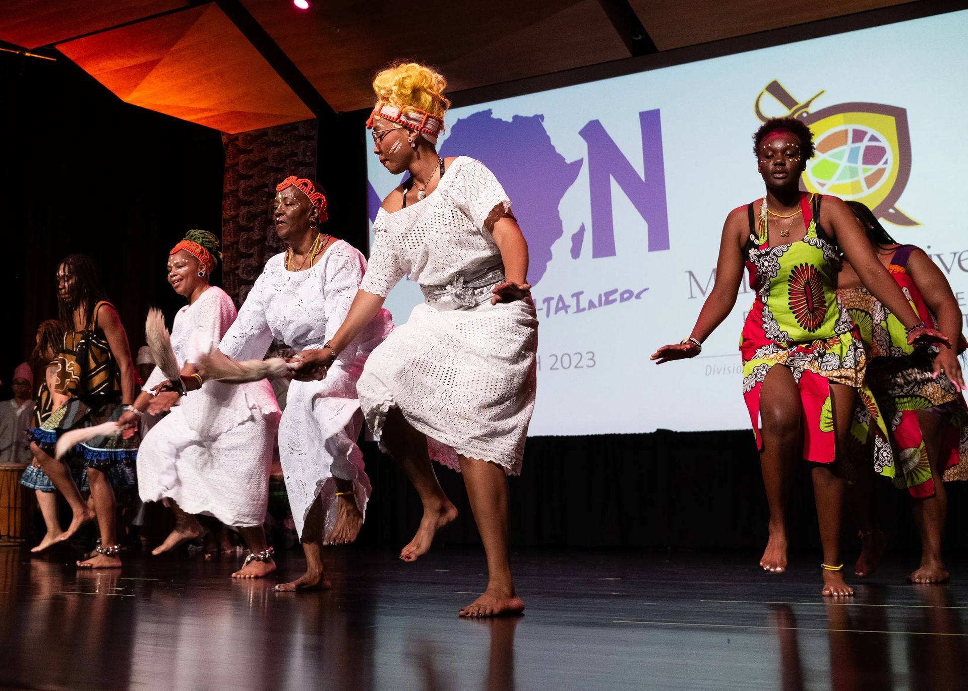 Women in colorful clothing dancing on stage. Backdrop with a logo that says 