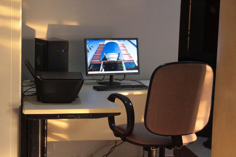Mesa com computador, roteador e cadeira em uma sala; a luz do sol ilumina o ambiente.