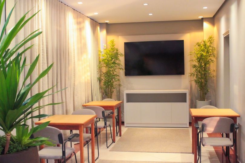 Sala pequena com TV, mesas, cadeiras, plantas e um tapete. Combinação de cores bege e marrom.