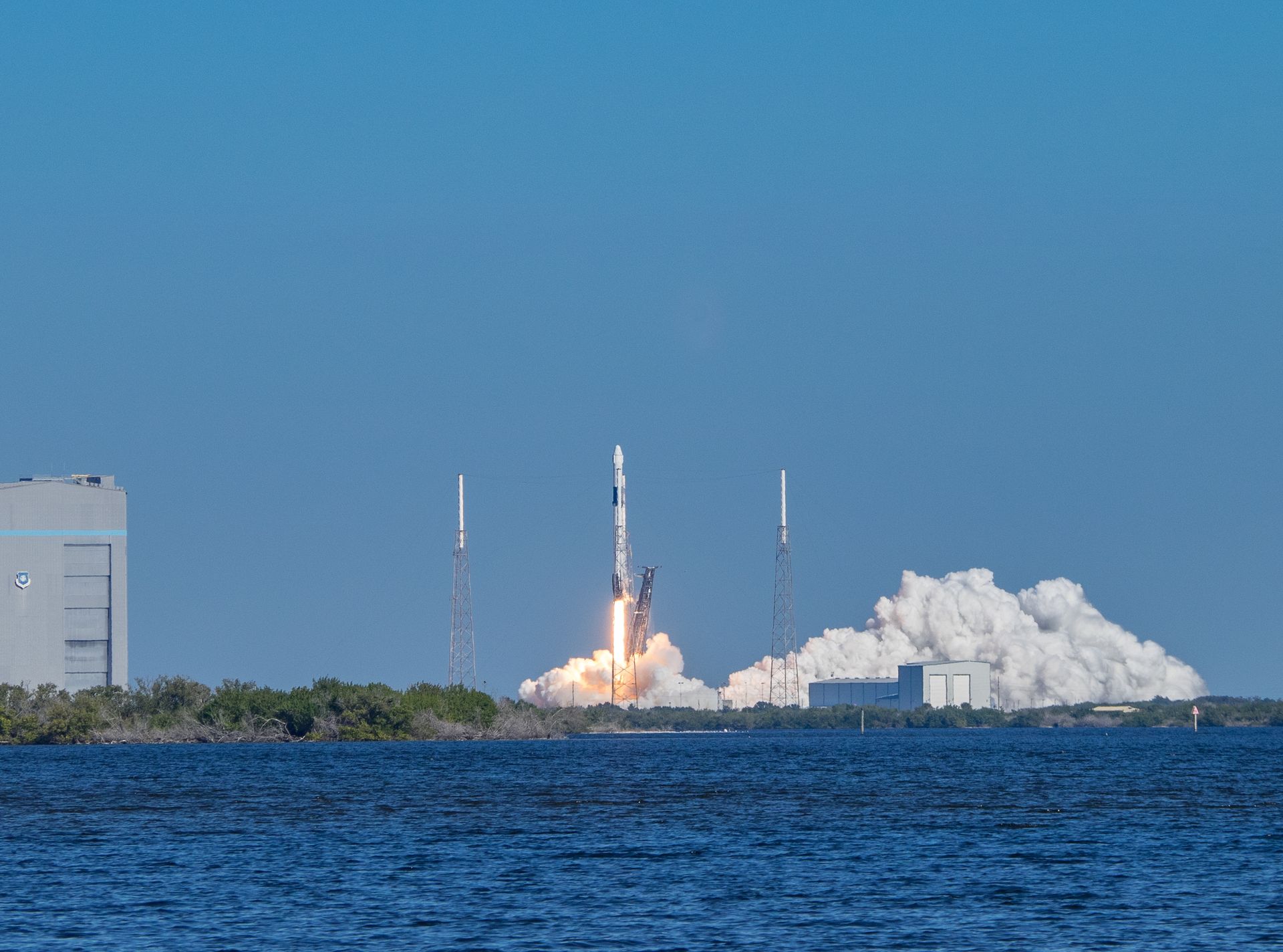 A rocket launches from a coastal site, emitting flames and smoke against a blue sky.