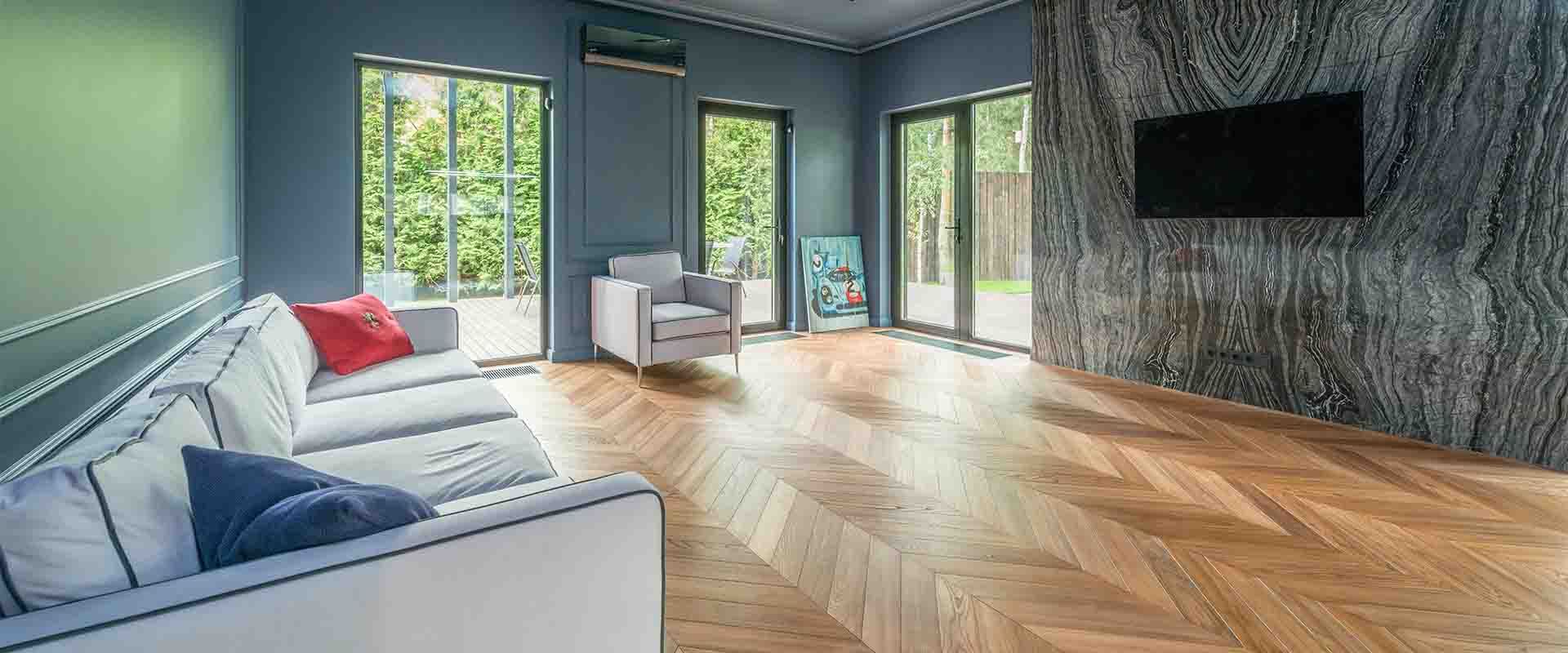 Modern living room with a light gray sofa, armchair, large television on a stone feature wall, and chevron wood flooring.