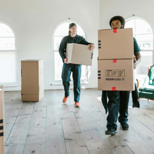 Two people in dark work uniforms carry cardboard moving boxes across a bright, empty room with wood floors and arched windows