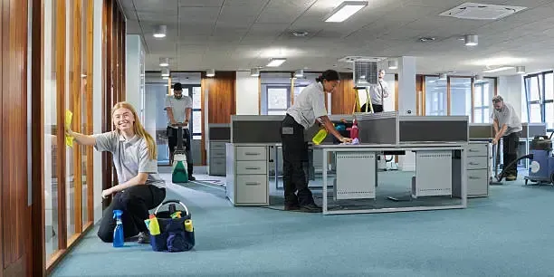 An open-plan office with beige cubicle partitions, several workstations, and two people working at their desks.