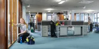 An open-plan office with beige cubicle partitions, several workstations, and two people working at their desks.