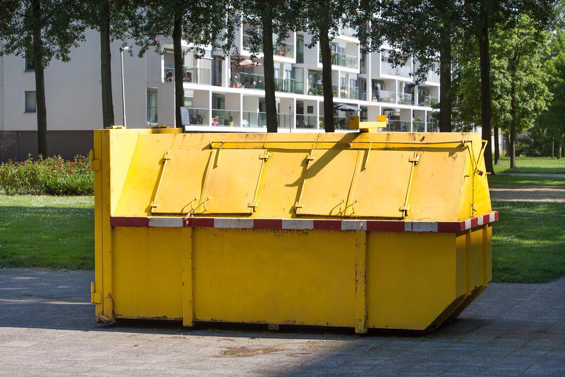 Yellow dumpster on a paved roadside with trees and an apartment building in the background