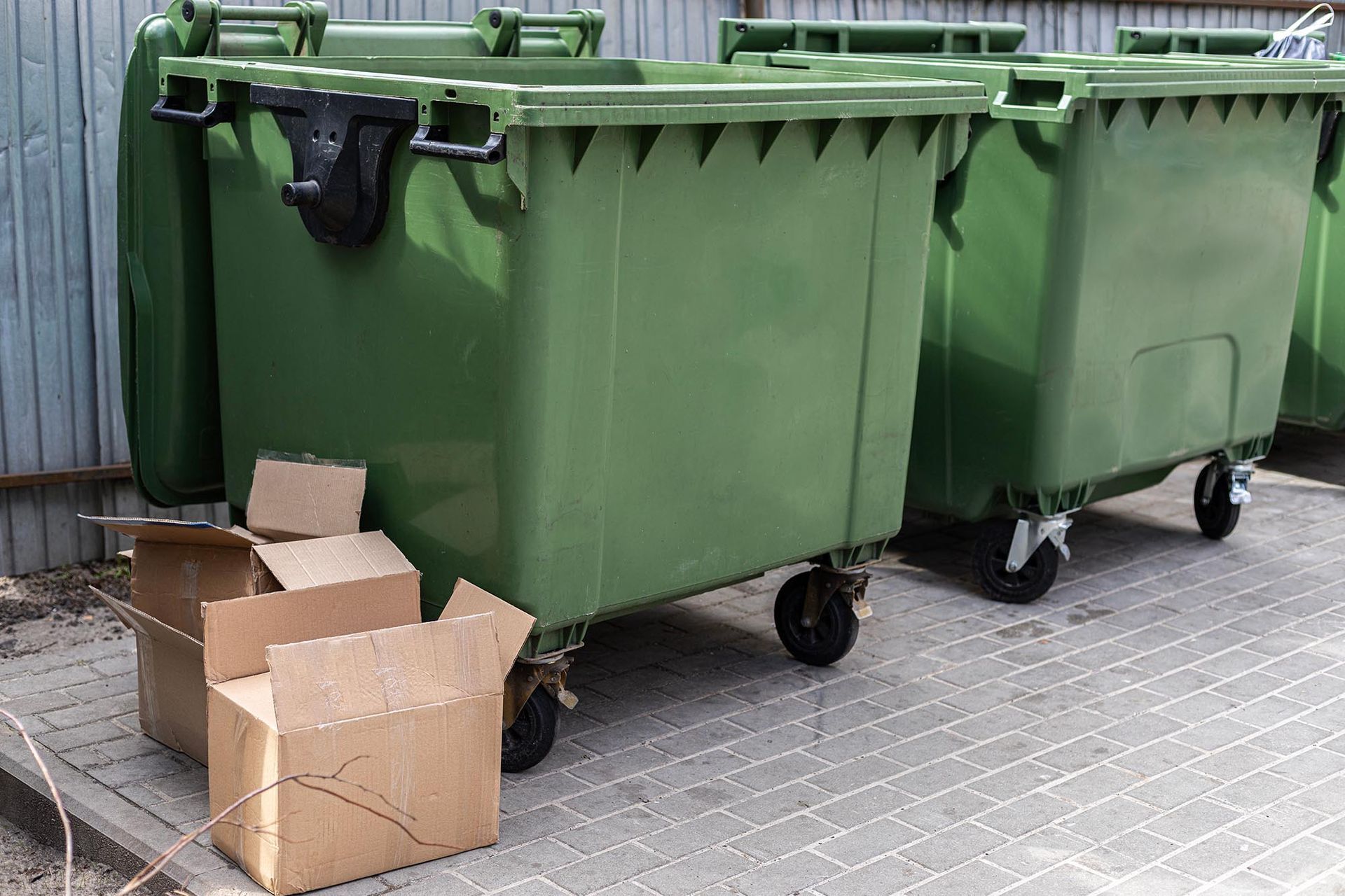 Green dumpsters beside several open cardboard boxes on a paved outdoor area