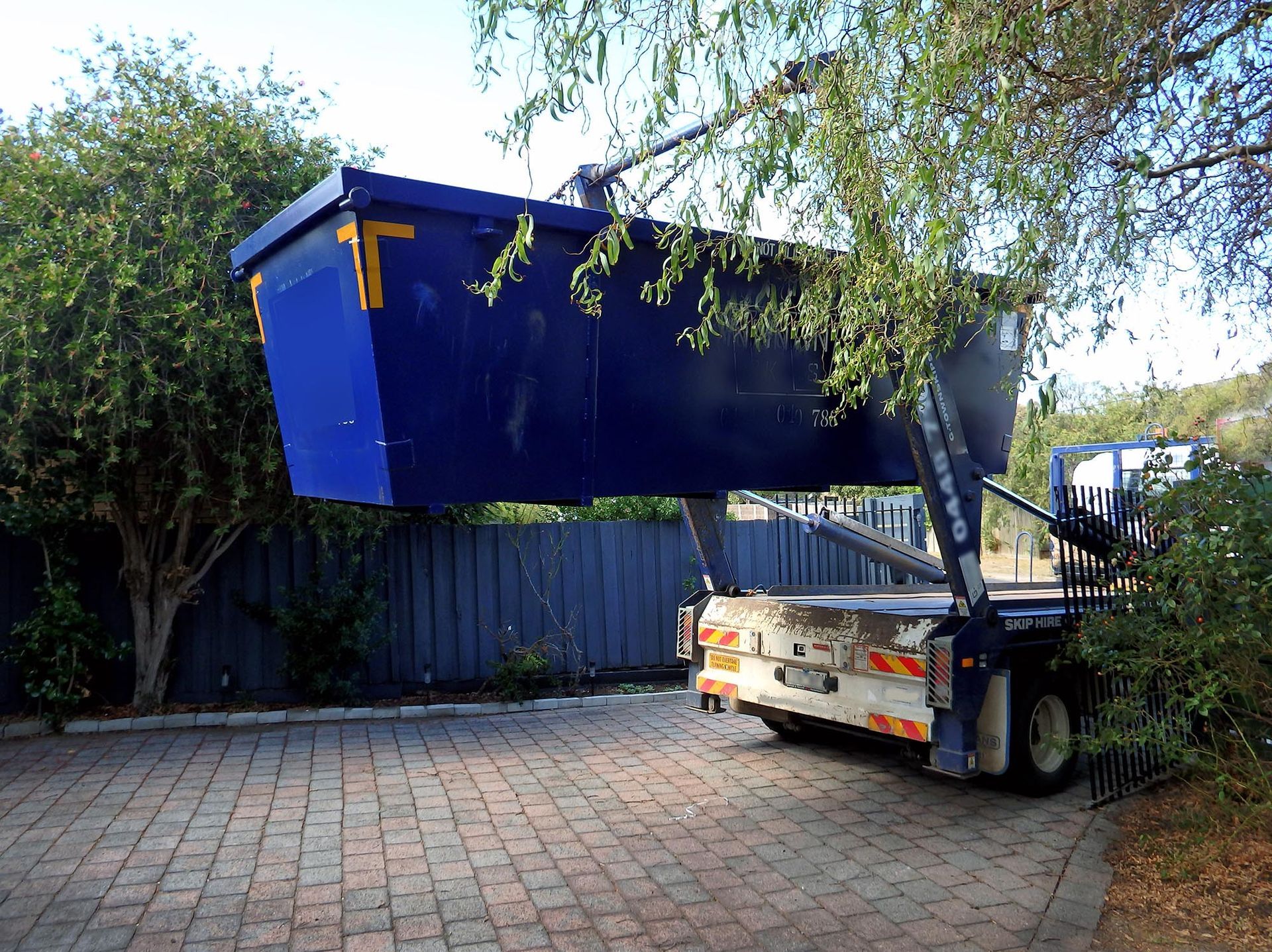 Blue dumpster being lifted by a white truck in a paved yard, with trees and a fence nearby.