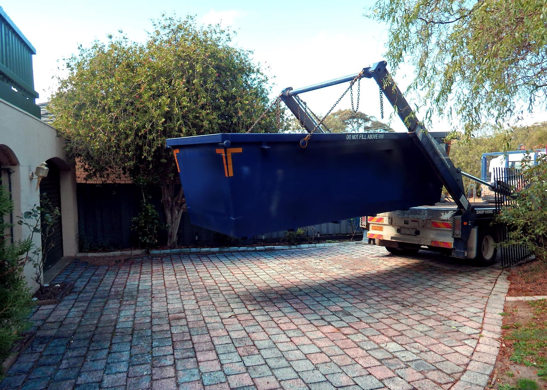 Small crane loading a large blue dumpster onto a truck in a paved driveway.