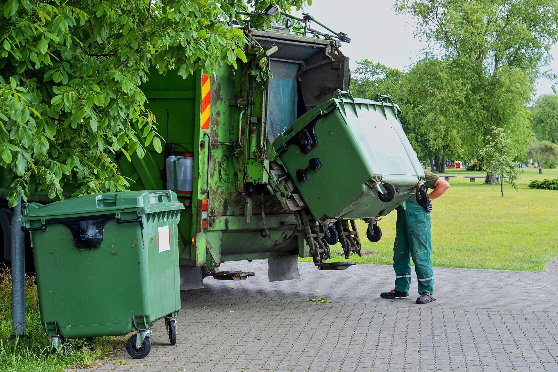 Garbage truck unloading green trash bins on a paved street beside trees and grass