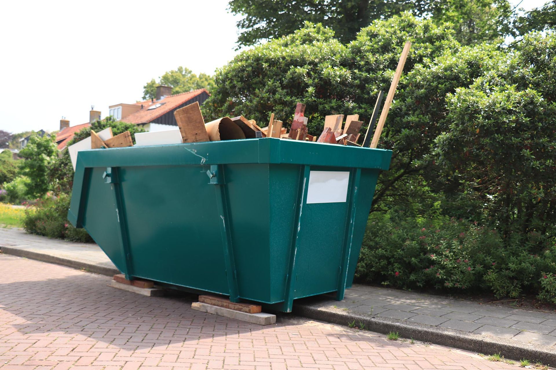 Large green dumpster filled with wood debris on a sidewalk beside bushes and houses.