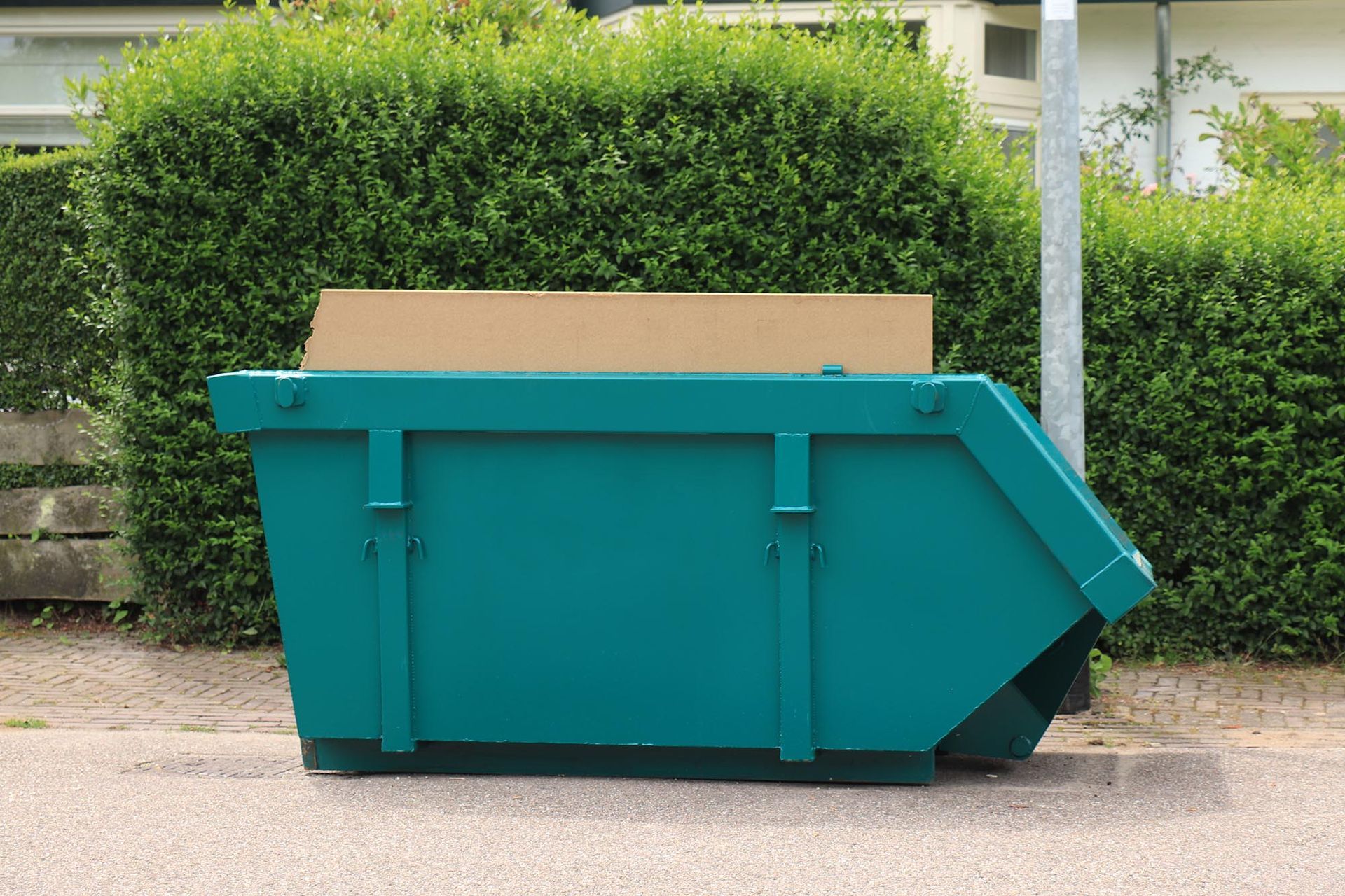 Teal construction dumpster on a street with green hedges behind it