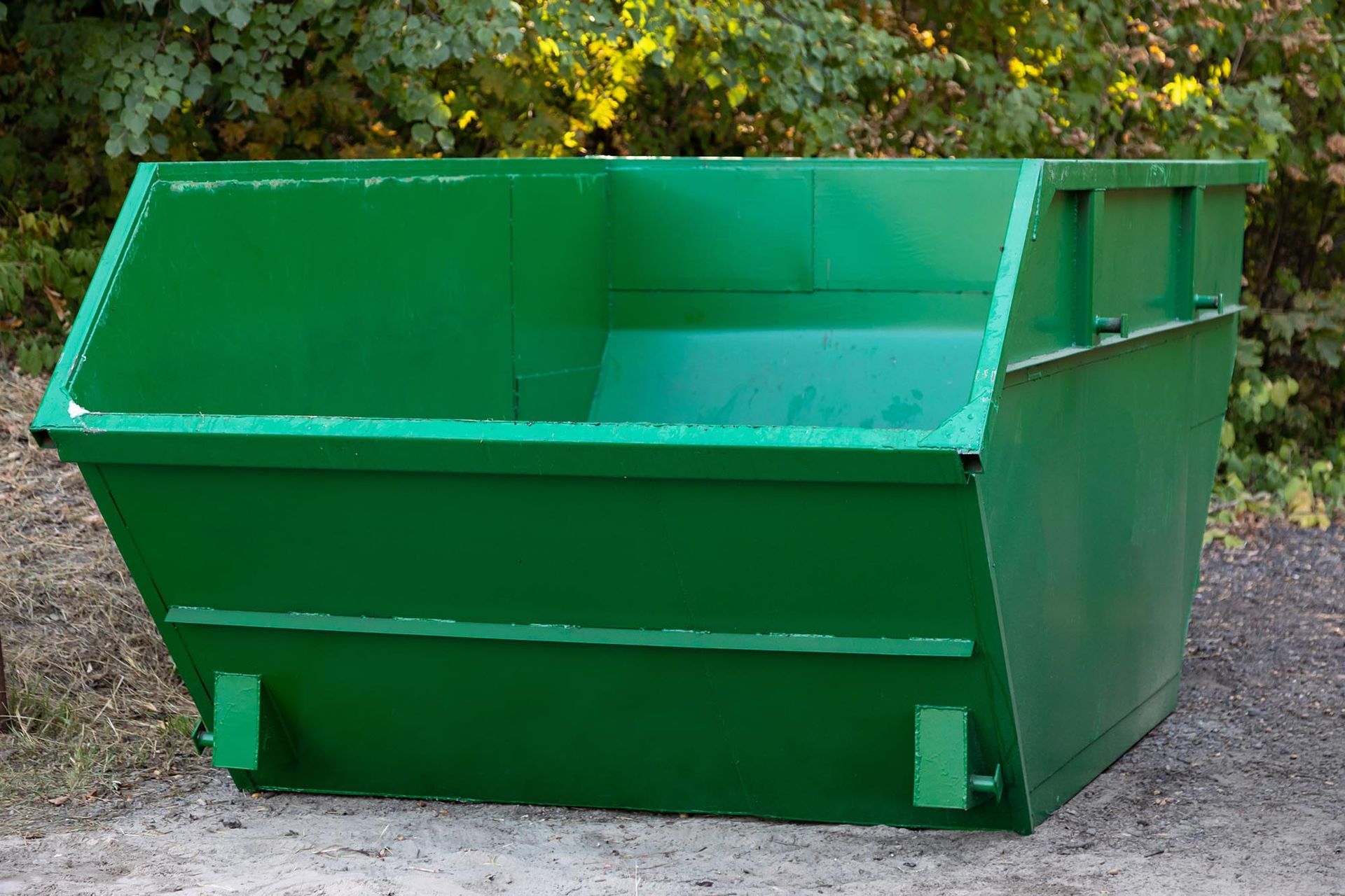 Green metal dumpster outdoors on gravel with trees in the background