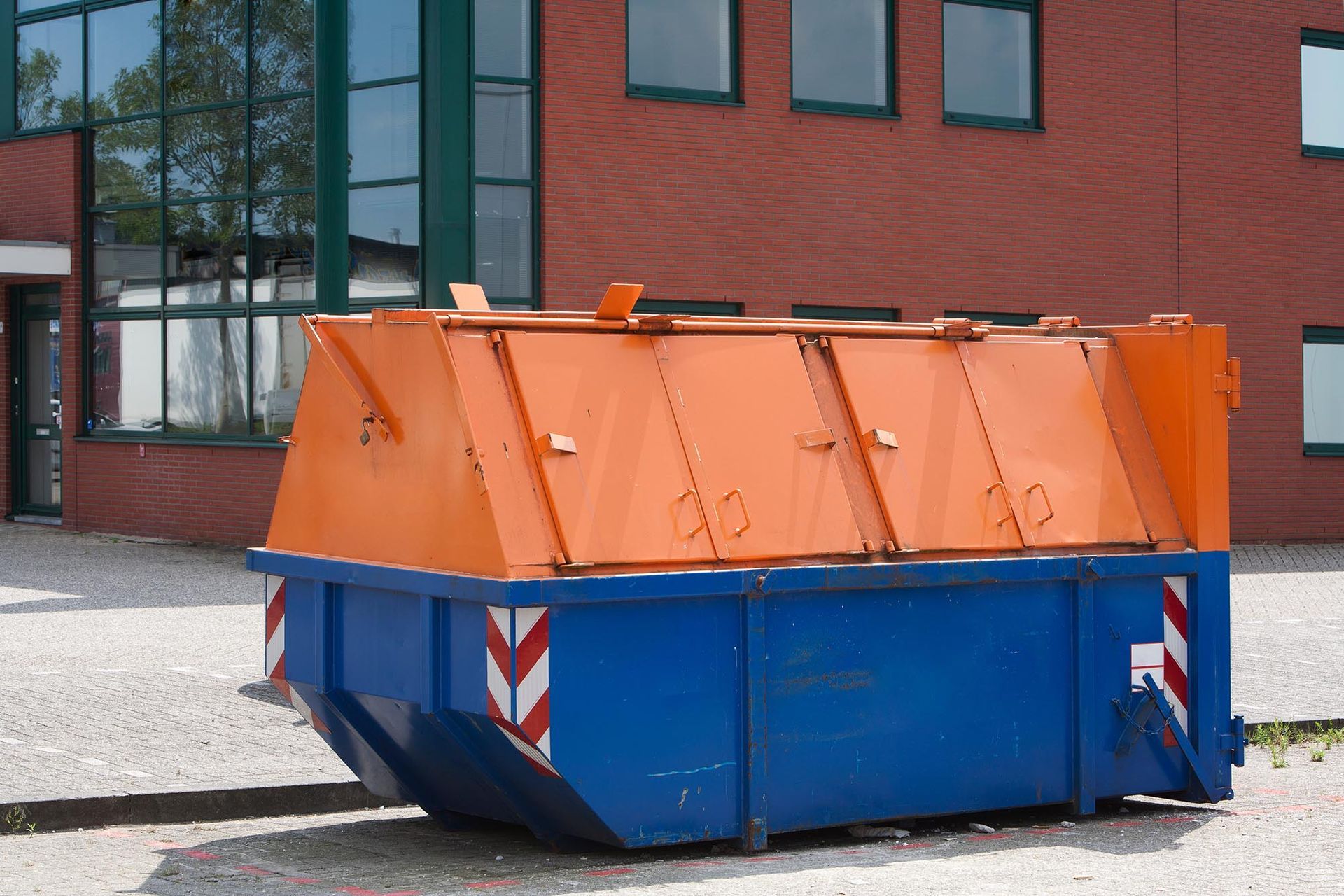 Large blue-and-orange industrial dumpster on a gravel lot beside a brick building