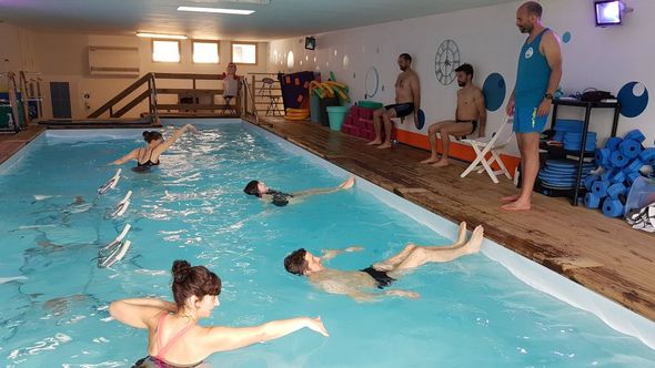Cours de natation en piscine intérieure avec des personnes qui font des longueurs et un instructeur debout au bord du bassin.
