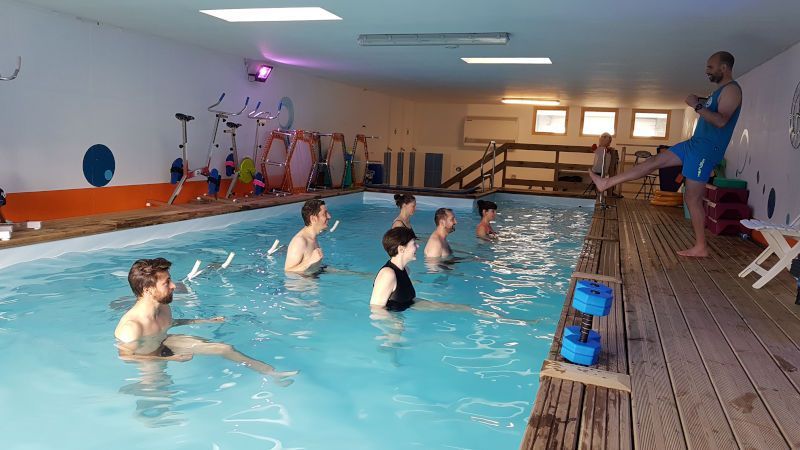Cours en piscine intérieure avec plusieurs personnes pratiquant dans l'eau tandis qu'un moniteur se tient sur le bord.