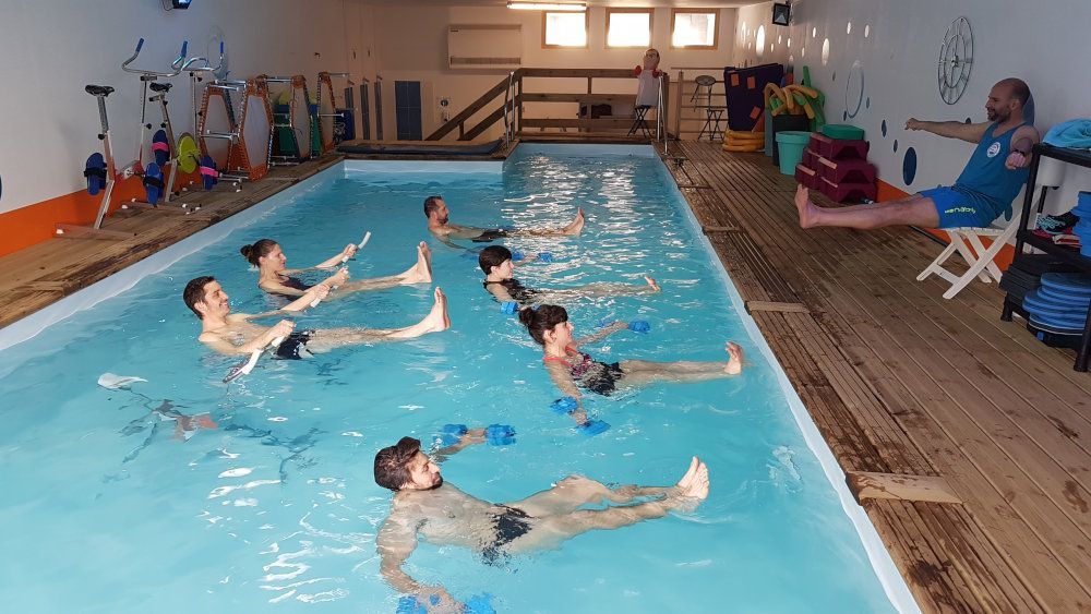 Des personnes font de l'aquagym dans une piscine intérieure, avec des moniteurs sur le bord du bassin.
