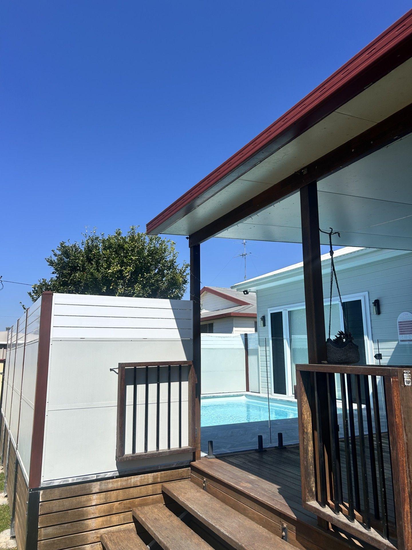 Outdoor deck with steps, leading to a fenced-in area with a swimming pool and a light blue house under a clear sky — Clarence Valley Pools In Grafton, NSW