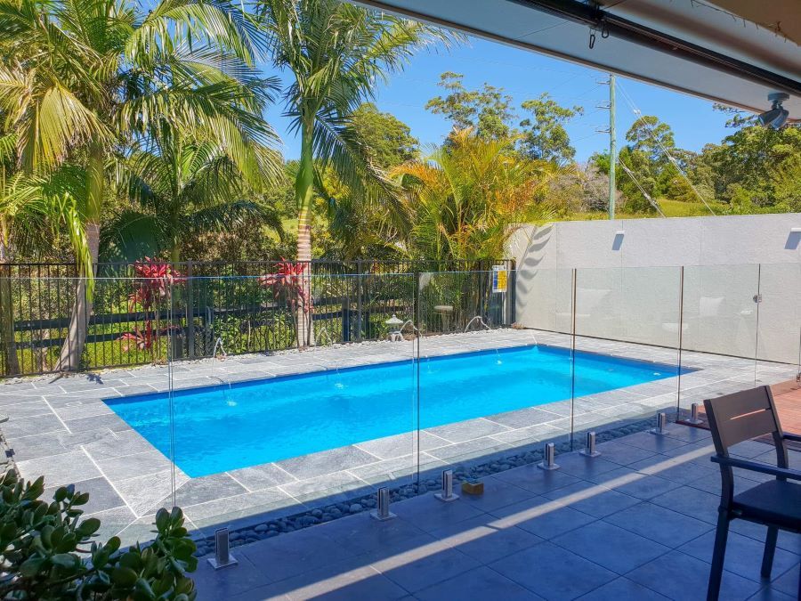 A Large Swimming Pool is Surrounded by a Glass Fence and Palm Trees — Clarence Valley Pools In Woolgoolga, NSW