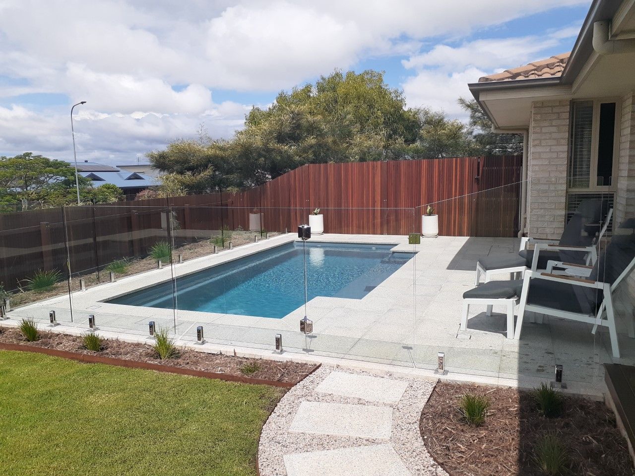 Swimming pool with glass fence, wooden fence, and stone pathway. Blue water, green lawn, sunny day— Clarence Valley Pools In Grafton, NSW
