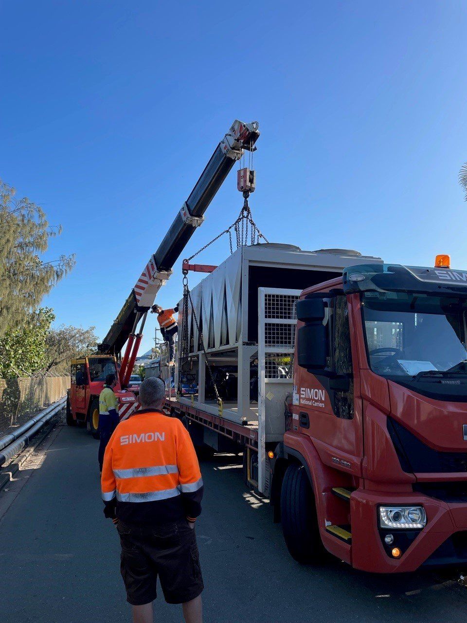 Crane lifting a large gray truck onto a red flatbed, with workers nearby on a roadside.